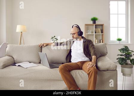 Mature man in headphones sitting on sofa at home and watching video on laptop computer Stock Photo