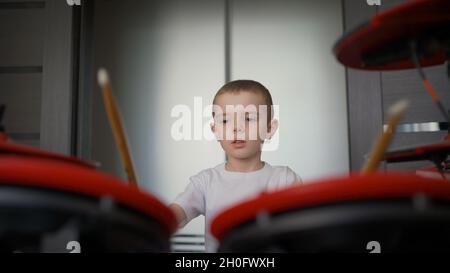 A little boy enjoys playing the electronic, red, drum at home. The boy at home on self-isolation exercises drums. A boy plays electronic drums at home Stock Photo