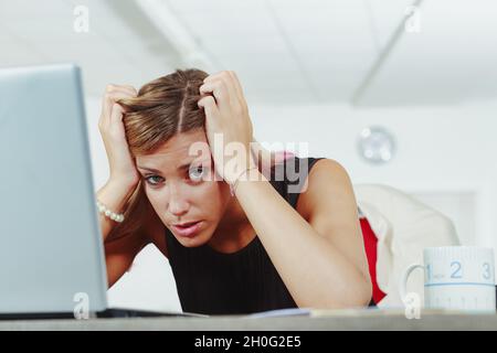 Frustrated stressed businesswoman with head in hands leaning over her laptop computer at her desk looking down at the camera Stock Photo
