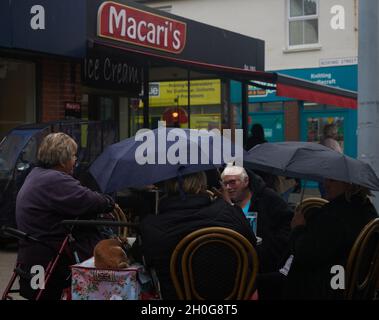 People enjoying their time together and having a drink outside on the terrace of Macaris café in Bognor Regis shielding from the rain with umbrellas. Stock Photo
