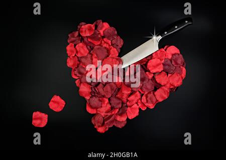 Red petals shaped into a heart with a knife sticking in with loose petals representing drops of blood, taken on a plain white background