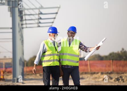 Two enthusiastic engineers with helmets and vests talking at building site in front of metal construction Stock Photo