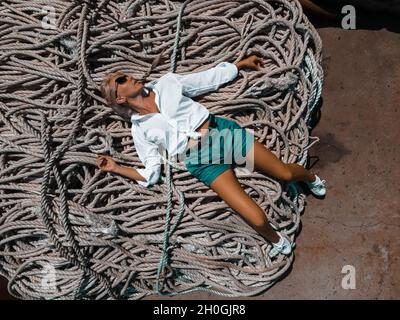 A young girl in a white shirt and blue shorts and sunglasses is lying on a pile of sea ropes. A drone photo up close. Stock Photo