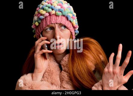 Confused young redhead girl with cap and jacket talking on phone Stock Photo