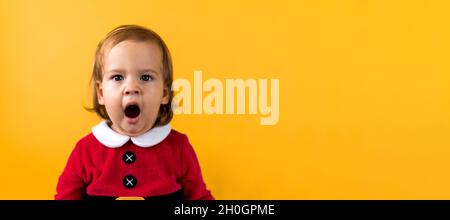 Banner Portraite Emotion Cute Cheerful Chubby Baby Girl in Santa Suit Yawns As If Singing At Orange Background. Child Christmas Scene Celebrating Stock Photo