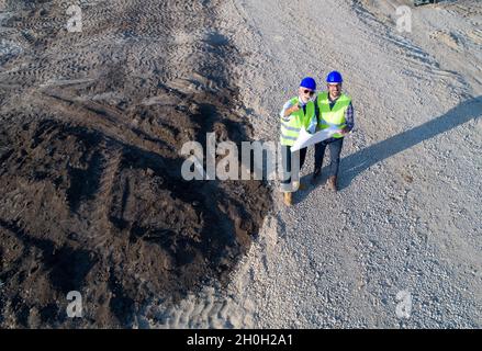 Aerial image of two engineers with helmets and vests, holding blueprints on construction site and looking up at drone Stock Photo