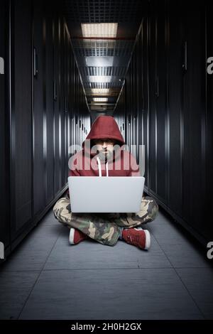 The hacker in a hood sits with the computer among servers, the burglar. Storage of data, cloudy services, system breaking, attack of hackers Stock Photo