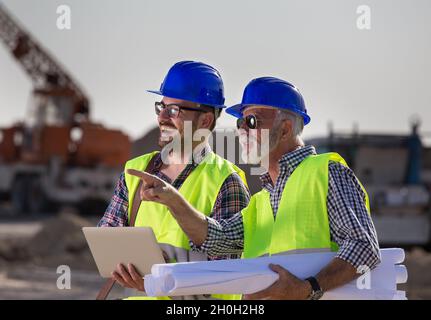 Twp satisfied engineers talking at building site with construction machinery in background Stock Photo