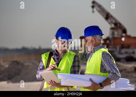 Two satisfied engineers talking at building site with construction machinery in background Stock Photo