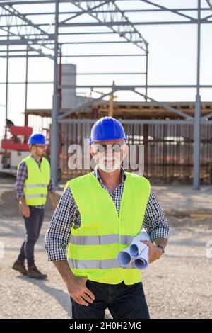 Senior engineer holding blueprints in front of metal structure on building site Stock Photo