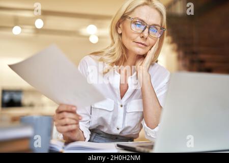 Focused mature businesswoman discussing papers during online video conference using laptop while sitting at her desk in the office Stock Photo