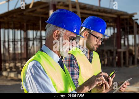 Senior engineer and worker looking at cell phone and tablet in front of construction at building site Stock Photo