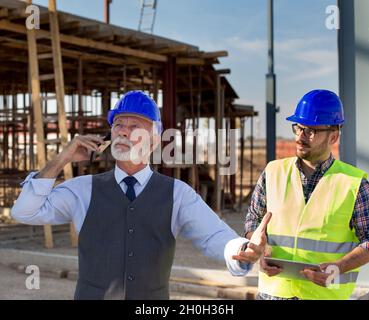 Angry senior engineer with helmet talking on phone standing beside young worker at building site Stock Photo
