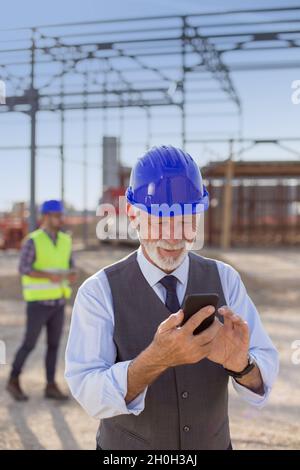 Senior engineer with helmet typing on mobile phone at building site Stock Photo