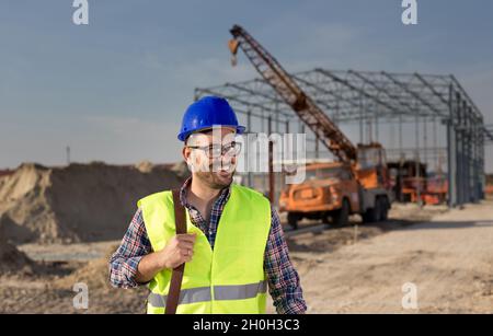 Happy young engineer with helmet standing in front of metal construction at building site Stock Photo