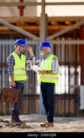 Two engineers with helmets and vests talking at building site in front of metal construction Stock Photo