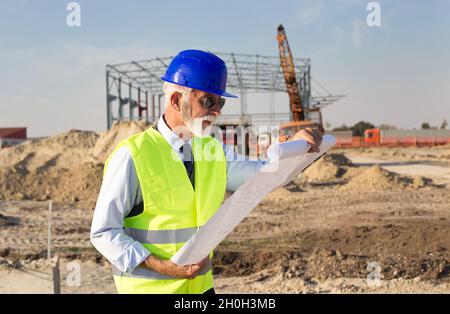 Senior engineer looking at blueprints in front of metal construction at building site Stock Photo