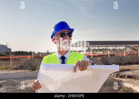 Senior engineer with blueprints standing at building site and looking up in the air Stock Photo