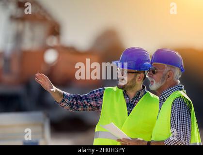 Two satisfied engineers talking at building site with construction machinery in background Stock Photo