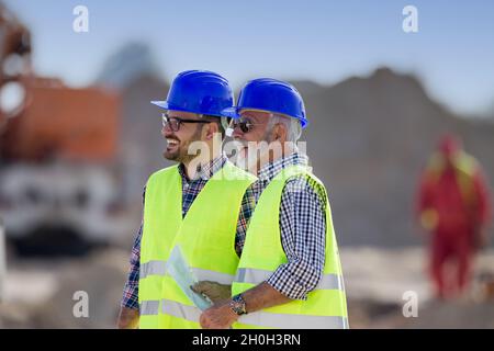 Two satisfied engineers talking at building site with construction machinery in background Stock Photo