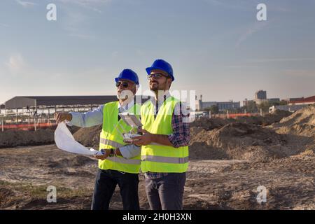 Two engineers with helmets and vests operating with drone by remote control and looking up in the sky. Technology innovations in construction industry Stock Photo