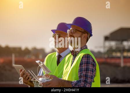 Two engineers with helmets and vests operating with drone by remote control and looking up in the sky. Technology innovations in construction industry Stock Photo