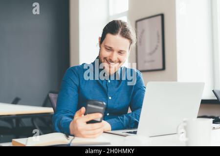 Smiling young male office worker using smartphone while working on laptop in modern office centre Stock Photo