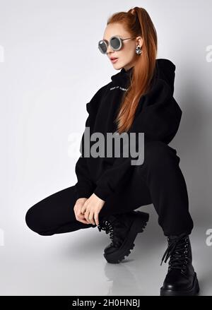 Slim fashionable redhead woman in trendy black sportswear hoodie, pants and massive brutal shoes sits squatted sideways Stock Photo