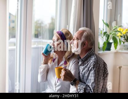 Happy senior couple standing beside window and drinking tea Stock Photo