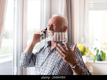 Excited old man in shirt with white hair and beard talking on phone beside window at home Stock Photo