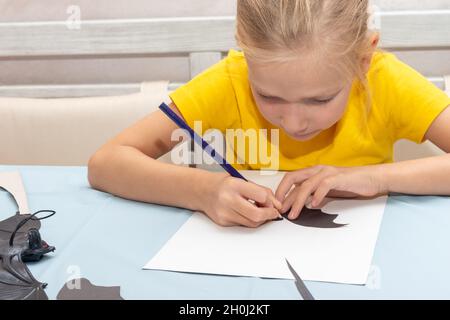 A girl draws and cuts a Halloween decoration at home from black paper. DIY craft decorations. Child makes black bats out of paper, origami Stock Photo