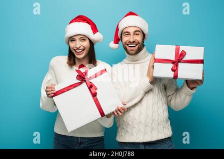 joyful couple in white sweaters and santa hats holding presents and looking at camera on blue Stock Photo