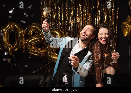 elegant and cheerful couple with champagne glasses and sparklers near golden christmas decoration on black Stock Photo