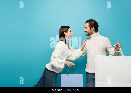 joyful couple in winter sweaters holding shopping bags and looking at each other on blue Stock Photo
