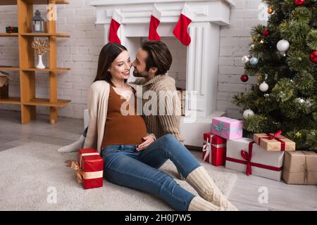 joyful pregnant woman with husband sitting on floor near decorated fireplace and presents under christmas tree Stock Photo