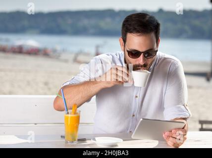 Handsome young man with sunglasses sitting in cafe on river beach, drinking coffee and looking at tablet Stock Photo