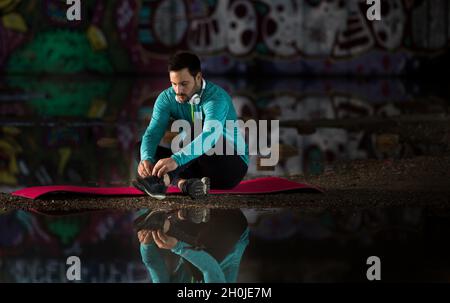 Handsome man sitting on mat and tying laces beside water with reflection of graffiti wall. Urban underground workout lifestyle Stock Photo
