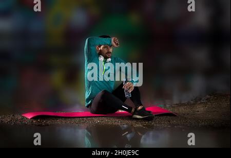 Tired man sitting on yoga mat, holding bottle of water and wiping sweat from forehead Stock Photo