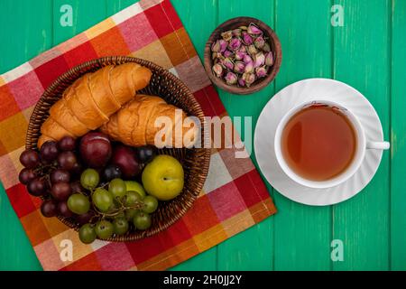 top view of fruits as grape pluots sloe berries with croissants in basket on plaid cloth and bowl of flowers with cup of tea on green background Stock Photo