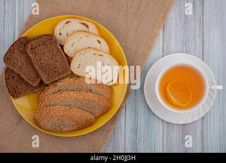 top view of breads as sliced seeded brown cob rye white ones in plate on sackcloth and cup of hot toddy on wooden background Stock Photo