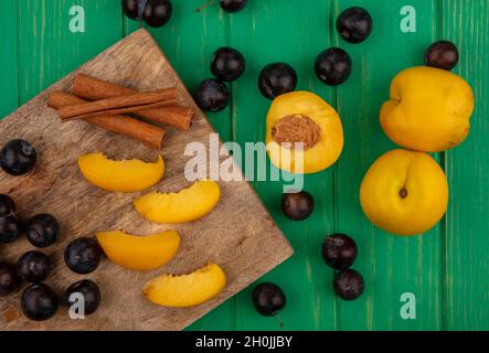 top view of fruits as sliced apricots and sloe berries with cinnamon on cutting board and half one with whole ones on green background Stock Photo