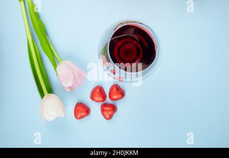top view of white and pink color tulip flowers with scattered heart shaped candies in red foil and a glass of wine on blue background