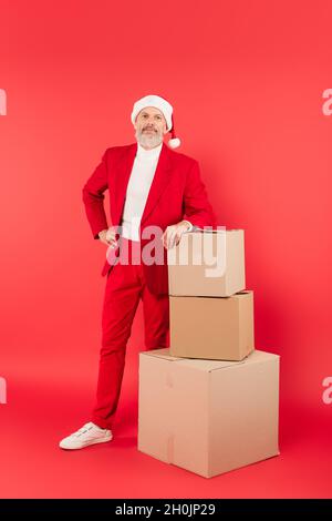 middle aged man in suit and santa hat leaning on carton boxes on red Stock Photo