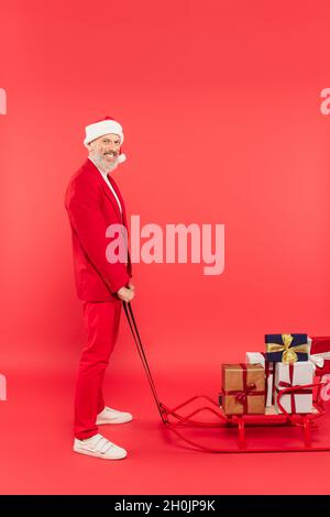 full length of cheerful middle aged man in santa hat and suit holding sleigh with presents on red Stock Photo