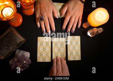 Detail of a woman's hands showing three tarot cards face down on a black card table, while another hand holds up one of the cards. Concept of divinati Stock Photo