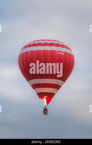 Single red and white hot air balloon in flight with flame, cloudy sky, people in wicker basket. Stock Photo