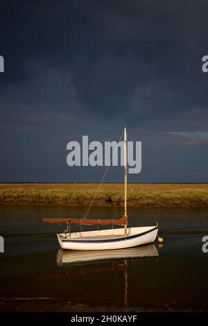 Late afternoon sun illuminates a small white sailing boat moored in Morston Creek on the north Norfolk Coast Path with dark sky before a storm Stock Photo