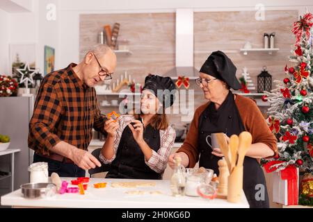 Happy family cooking traditional gingerbread dessert making homemade dough using baking tree shape in xmas decorated kitchen. Grandchild enjoying christmas holiday together preparing cookies Stock Photo