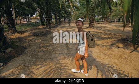 Woman tourist with plait walks looking around at growing young trees with lush leaves at oil palm farm elaeis guineensis on sunny day. Stock Photo