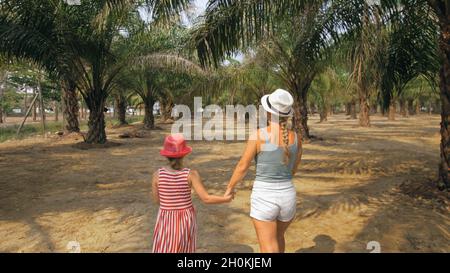 Mom and daughter for a walk. Woman tourist with plait walks looking around at growing young trees with lush leaves at oil palm farm elaeis guineensis Stock Photo